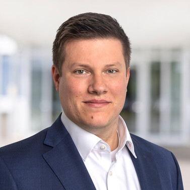 Porträt von Nico Pfeifer mit dunklem Sakko und weißem Hemd vor verschwommenem Bürohintergrund.